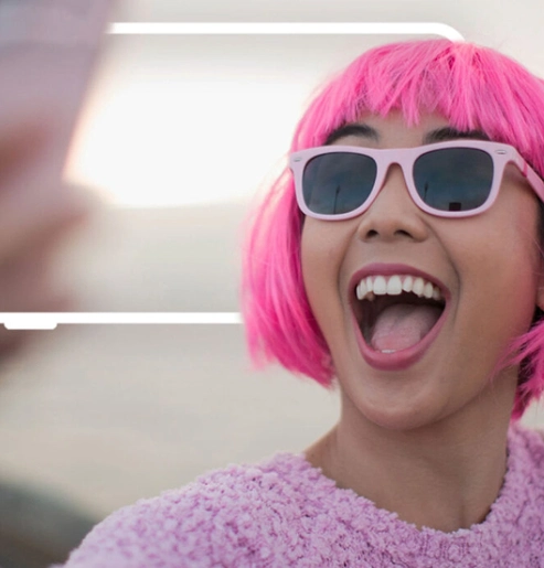 Una persona con el pelo corto de color rosa brillante y gafas de sol de montura rosa está riendo con la boca abierta mientras mira hacia un lado. Lleva un suéter de punto rosa.
