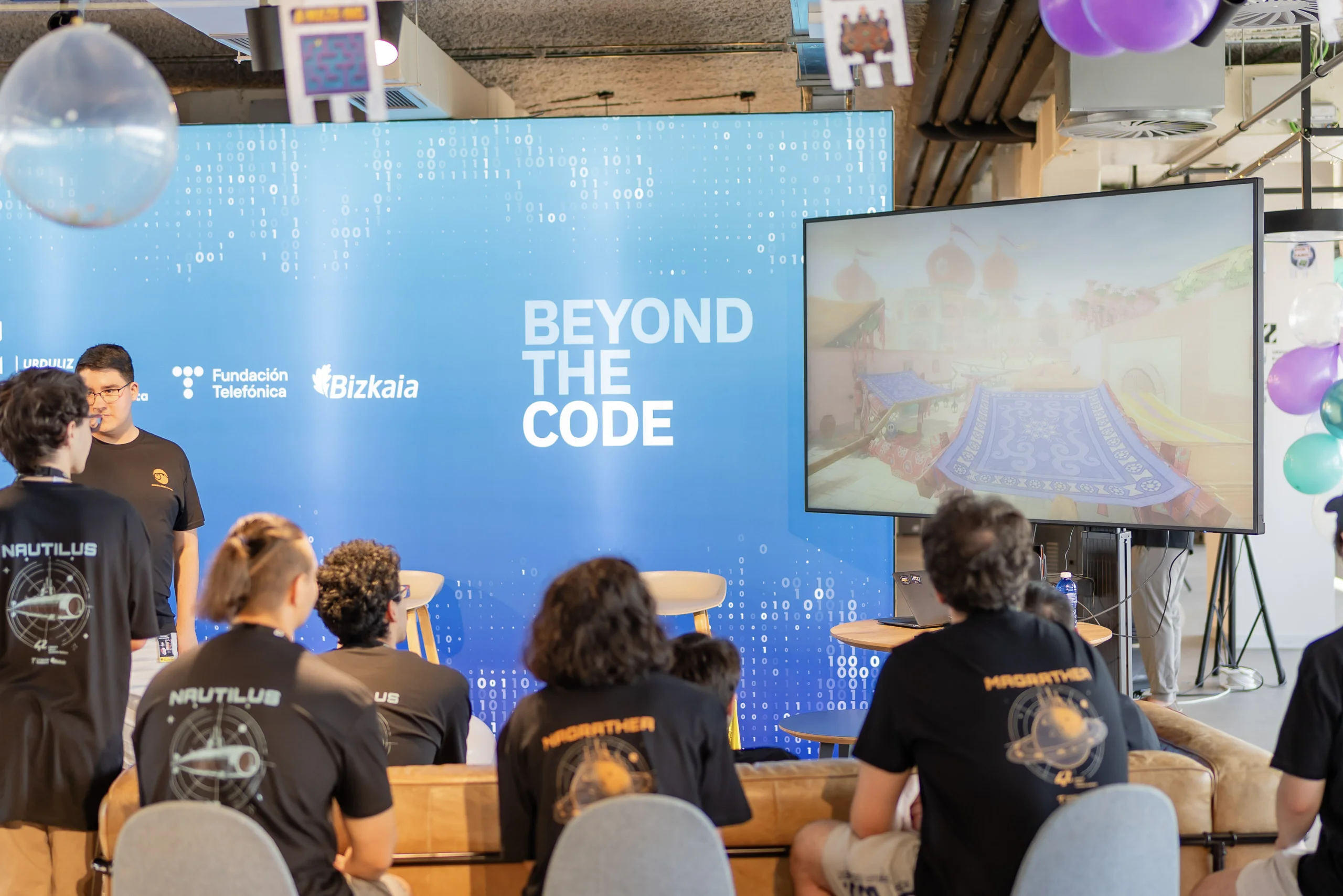 Público sentado en un evento tecnológico frente a un fondo azul en el que se lee «BEYOND THE CODE», con una gran pantalla a la derecha y globos colgando del techo.