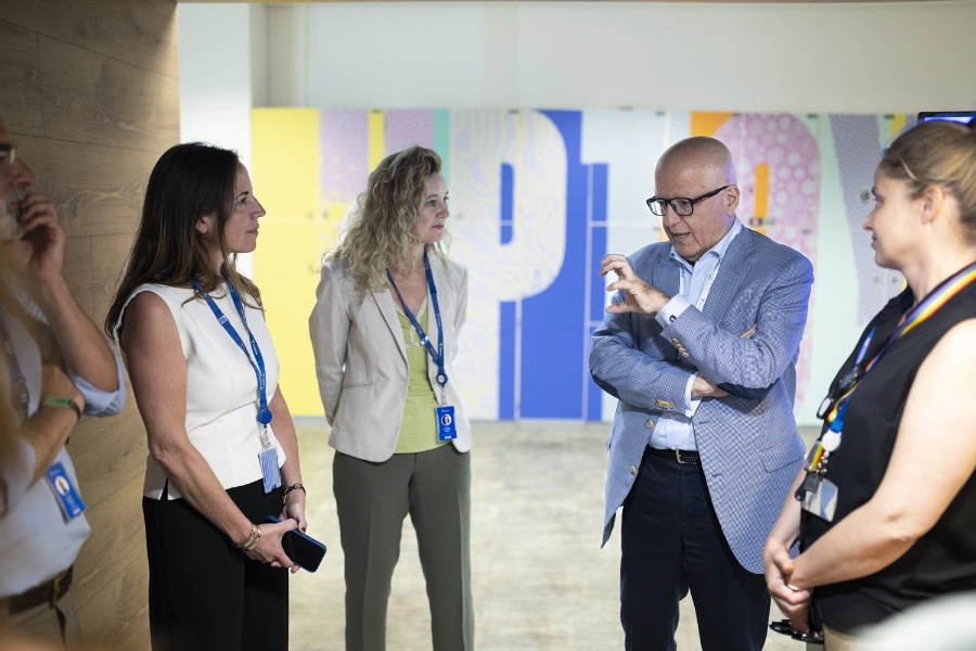 Un momento de la firma entre Fundación Telefónica y Fundación Pfizer para impulsar la salud digital de las personas mayores con Reconectados Salud Digital Five professionals in a hallway engaged in discussion; a man in a blue blazer gestures while others listen, colorful mural behind.