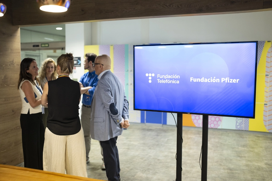 Un momento de la firma entre Fundación Telefónica y Fundación Pfizer para impulsar la salud digital de las personas mayores con Reconectados Salud Digital Group of professionals conversing near a large screen that reads Fundación Telefónica and Fundación Pfizer.