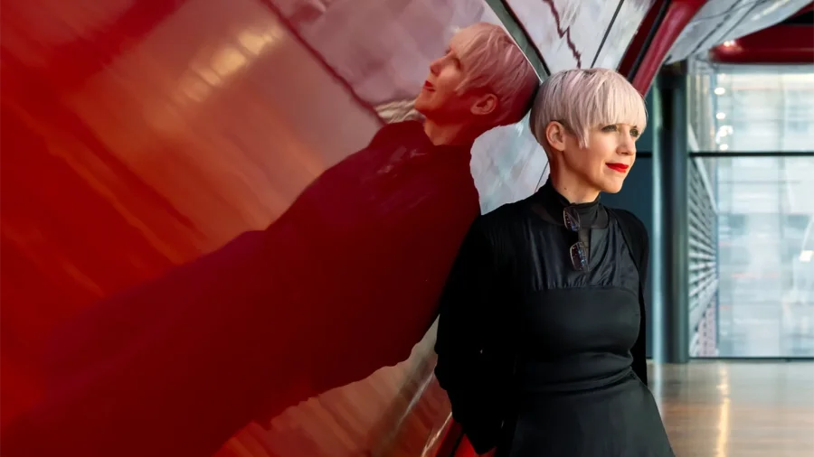 Two stylish women pose beside a shiny red wall in a modern hallway; the foreground woman wears a black dress and short platinum hair.