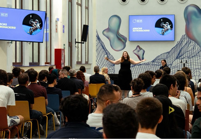 Imagen 1 A woman in a black dress stands with arms open on a stage addressing a seated audience, with two screens showing a space-themed slide behind her.