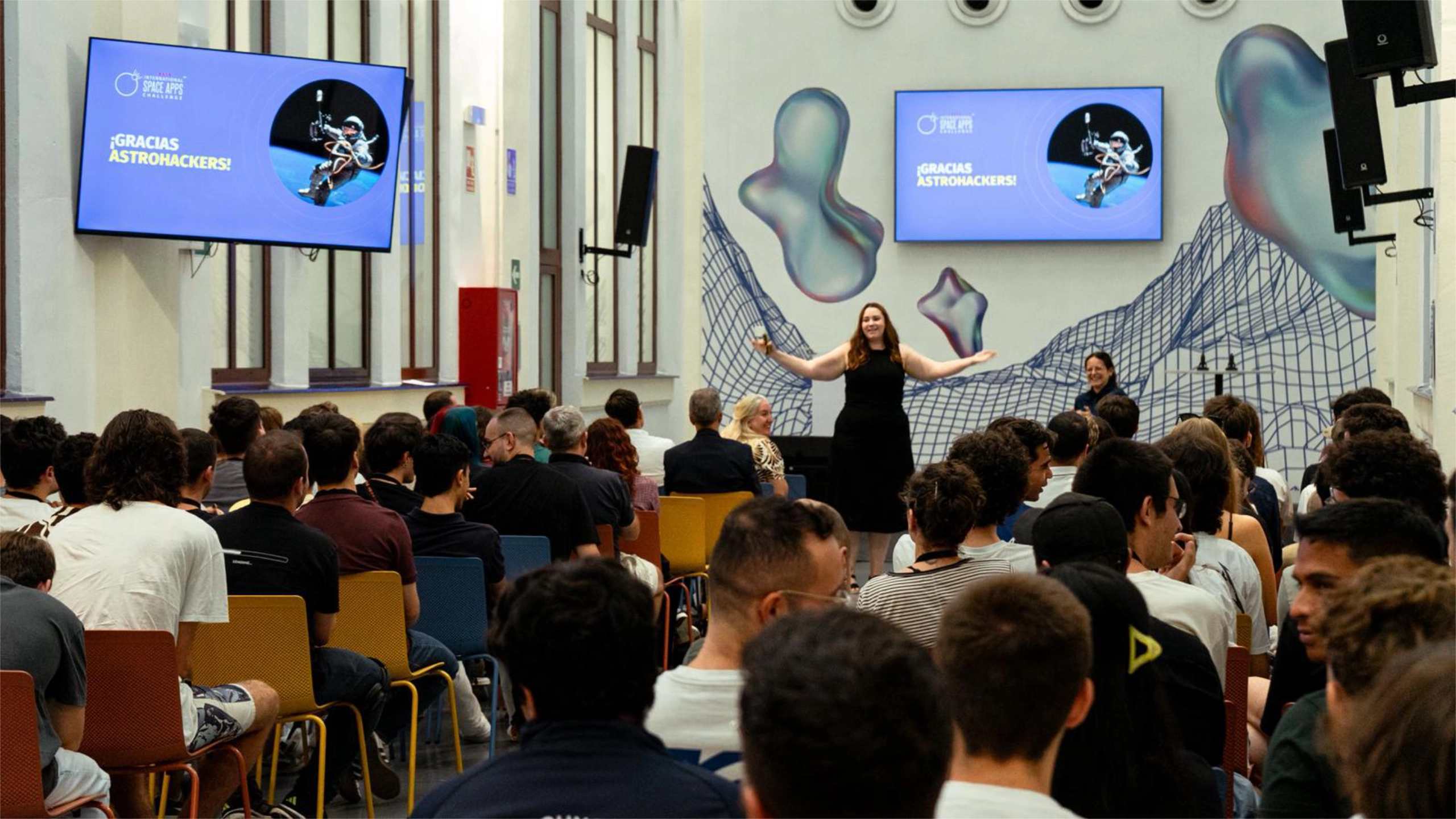 Presenter in a black dress speaks to a seated audience in a conference room, with two large screens showing 'Gracias ASTROHACKERS!' and blue graphics behind her.