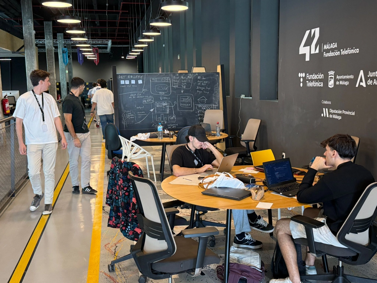 Open coworking space with a round table where two men work on laptops, a chalkboard wall in the background, and people walking along a yellow line.