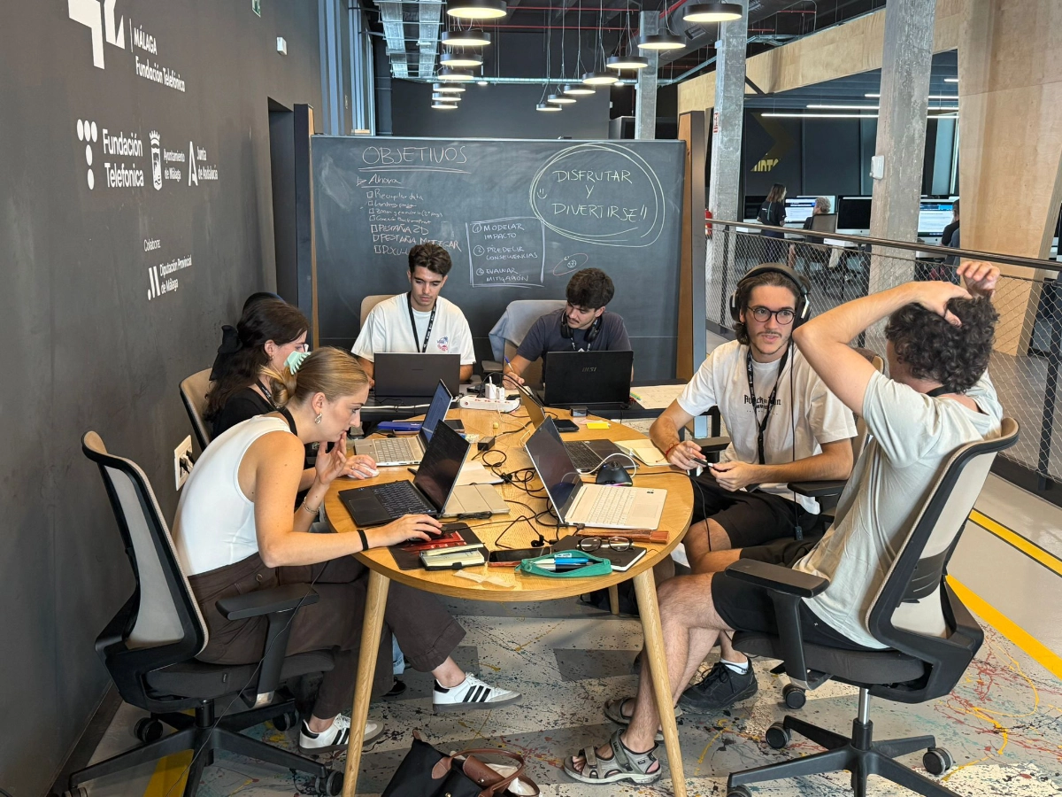Six people around a round wooden table with laptops and notebooks, collaborating in a modern office with a chalkboard reading 'OBJETIVOS' in the background.