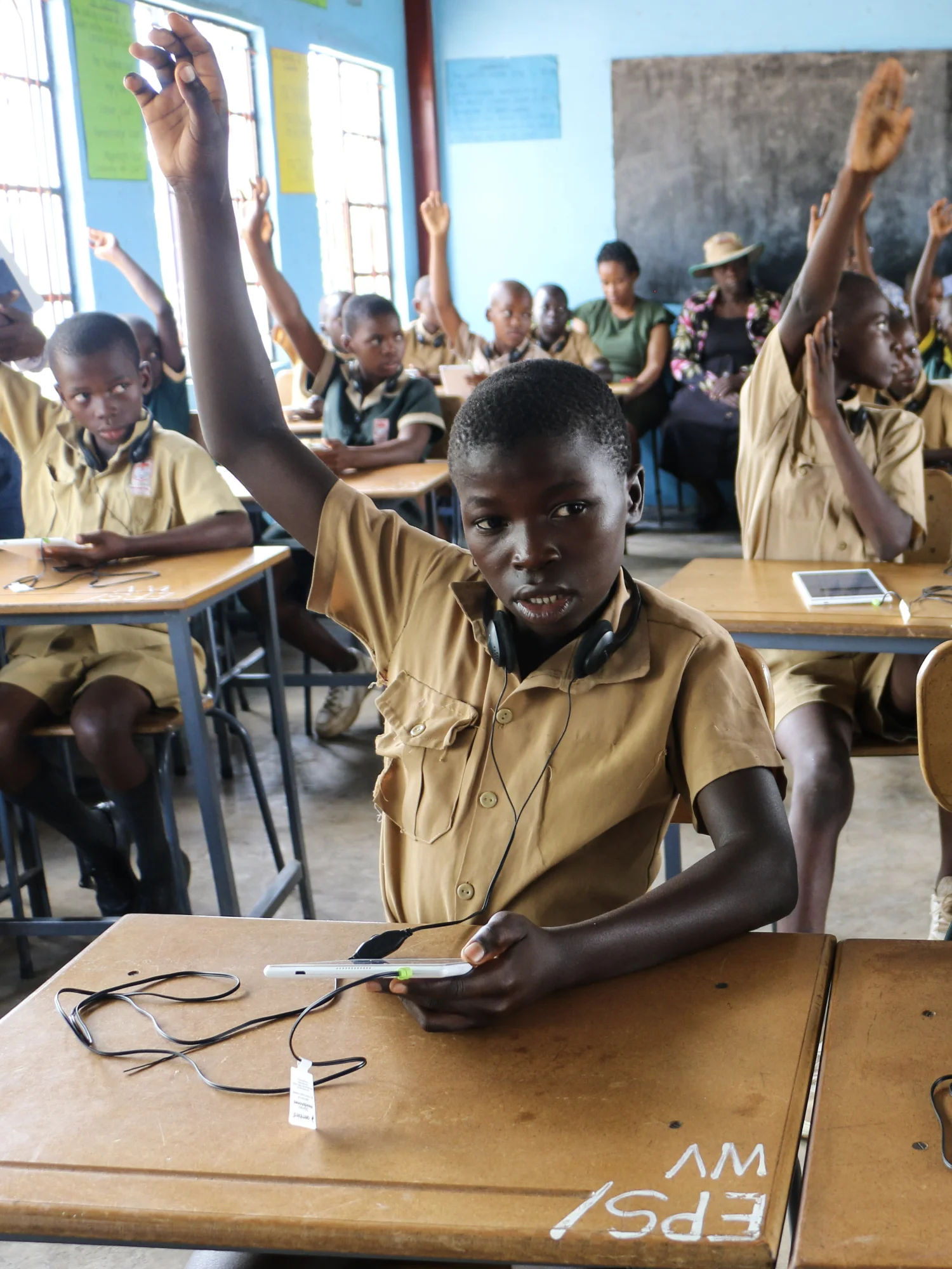 Un alumno levanta el brazo en el aula de un colegio Classroom of students in tan uniforms raising hands; foreground student with headphones sits at a desk with a phone and cables nearby.